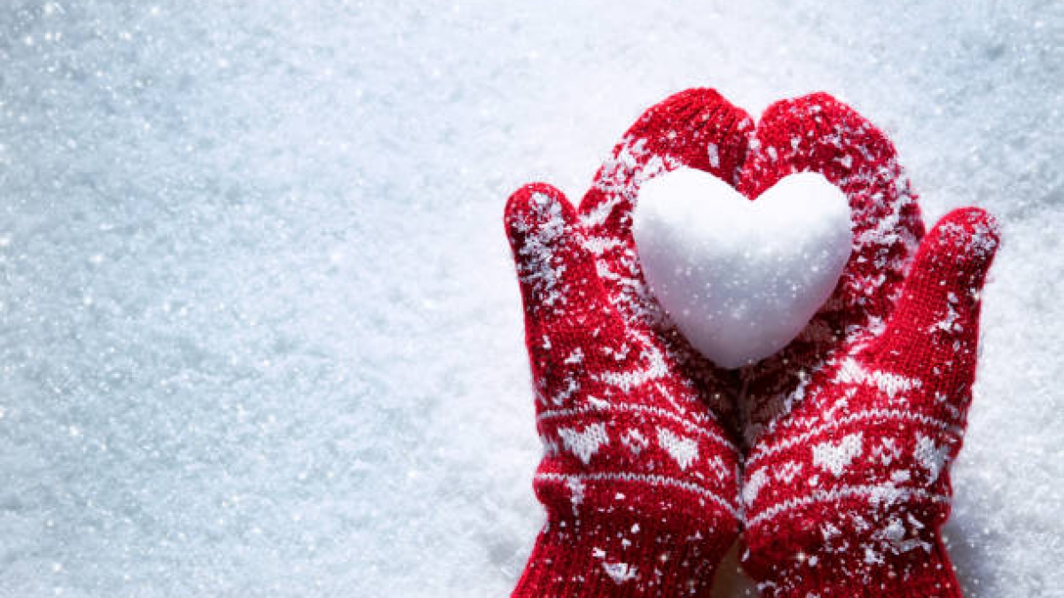 Female hands in knitted mittens with snowy heart against snow background. Love, winter and Valentines day romantic creative concept with copy space for text